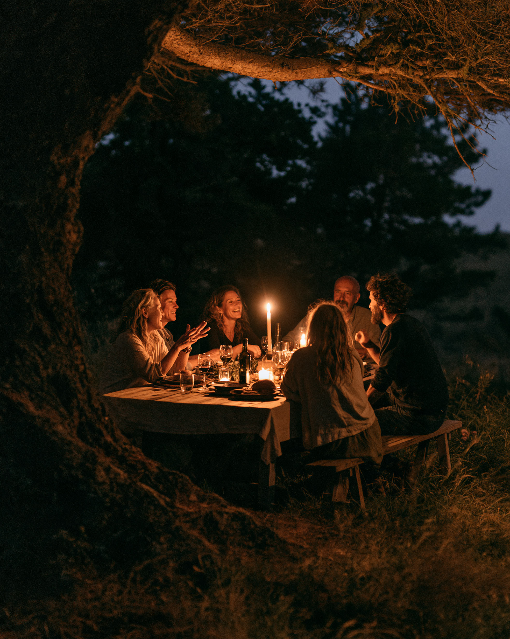 Friends gathered around a candlelit table under old trees at night