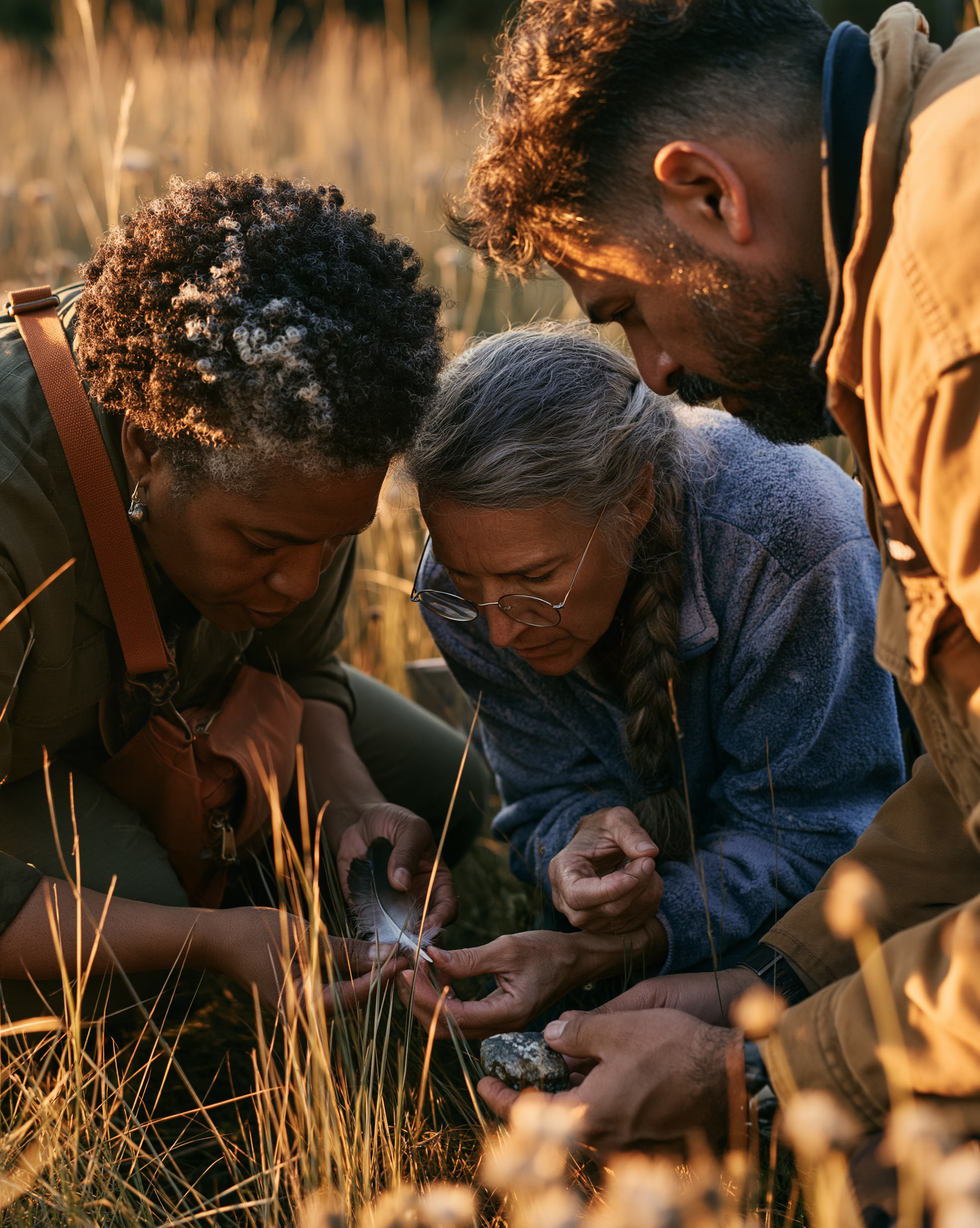 Three friends crouched in golden grass, examining a feather and a stone